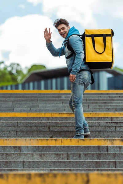Vista lateral do homem de entrega positiva com mochila térmica acenando a mão na câmera nas escadas — Fotografia de Stock