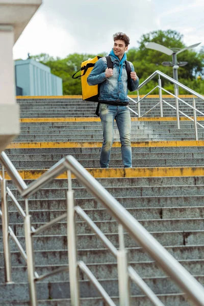 Selective focus of smiling courier with thermal backpack walking on stairs with railing — Stock Photo