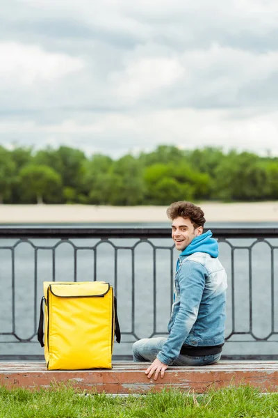 Back view of smiling courier sitting near thermo bag on bench on promenade — Stock Photo