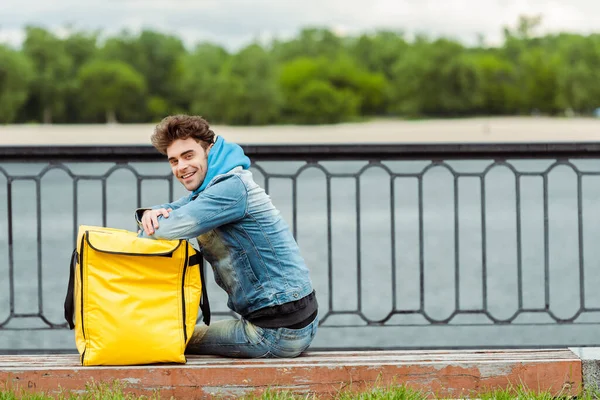 Side view of delivery man smiling at camera near thermo bag on bench on urban street — Stock Photo