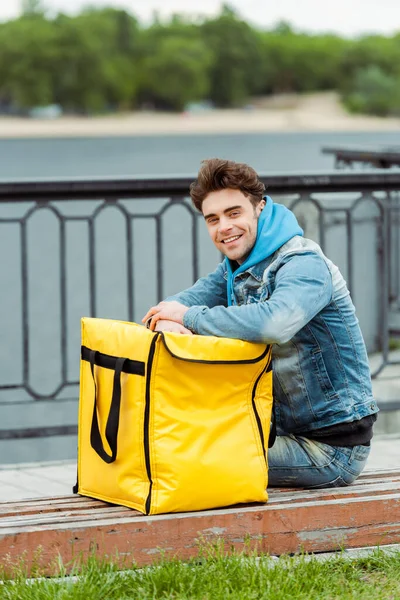 Side view of handsome courier smiling at camera near thermo bag on bench on urban street — Stock Photo