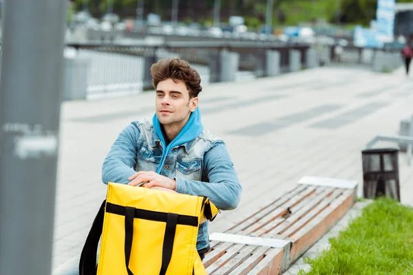 Selective focus of courier sitting near thermal bag on bench on urban street — Stock Photo