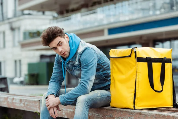Mensajero guapo mirando hacia otro lado mientras está sentado cerca de la bolsa térmica en el banco en la calle urbana - foto de stock