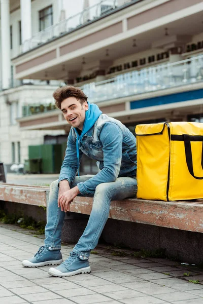 Handsome courier smiling at camera while sitting near thermo bag on bench on urban street — Stock Photo