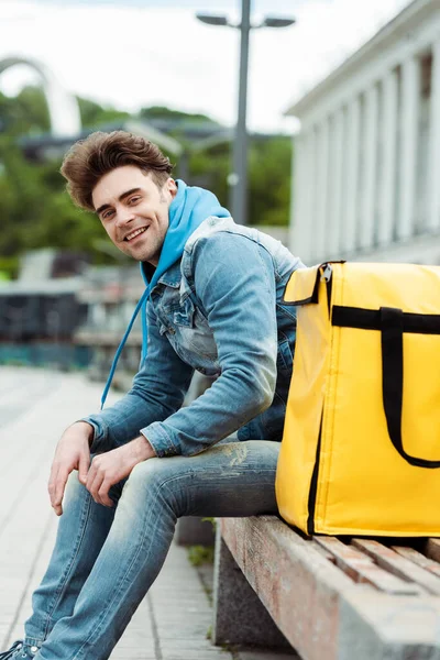 Selective focus of positive courier smiling at camera near thermo bag on bench on urban street — Stock Photo