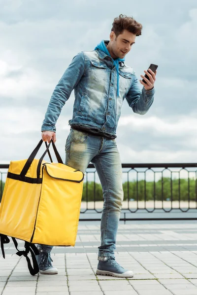 Mensajero sonriente con bolsa térmica mirando el teléfono inteligente en la calle urbana - foto de stock