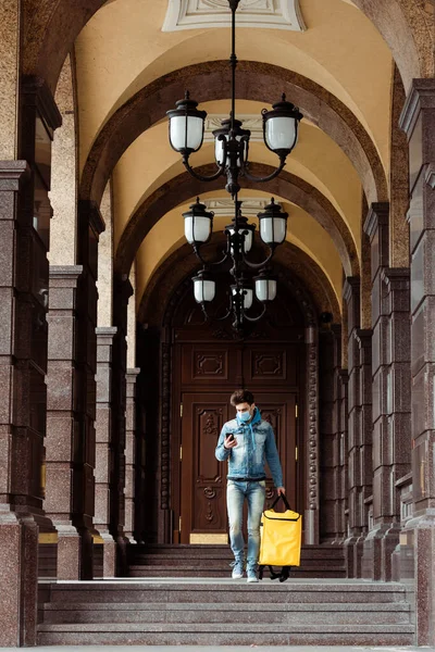 Доставщик в медицинской маске держит смартфон и тепловую сумку во время прогулки рядом со зданием с колоннами — стоковое фото
