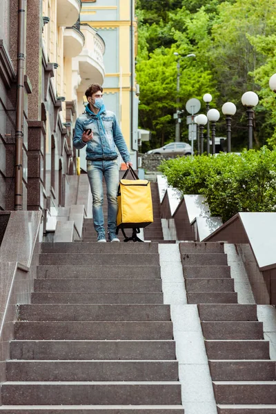 Courier in medical mask holding thermo bag and using smartphone on urban street — Stock Photo