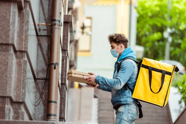 Side view of delivery man in medical mask and thermo backpack holding pizza boxes near building — Stock Photo
