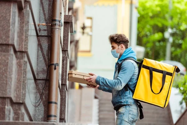 Vista laterale dell'uomo di consegna in maschera medica e zaino termo contenente scatole di pizza vicino all'edificio — Foto stock