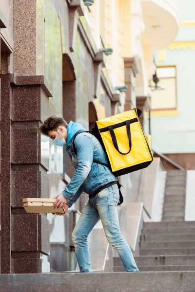 Side view of courier in medical mask and thermo backpack holding pizza boxes near building on urban street — Stock Photo