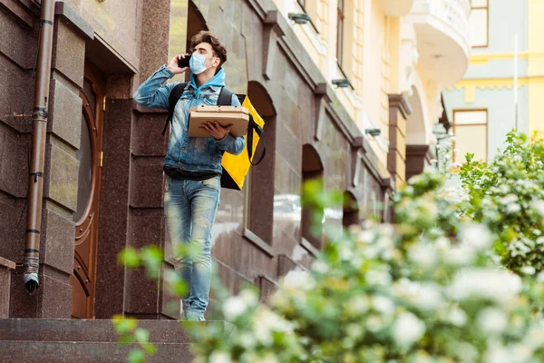 Foco seletivo do correio em máscara médica falando no smartphone e segurando caixas de pizza na rua urbana — Fotografia de Stock