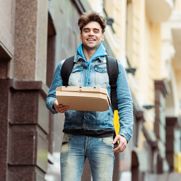 Mensajero guapo sonriendo a la cámara mientras sostiene cajas de pizza en la calle urbana - foto de stock