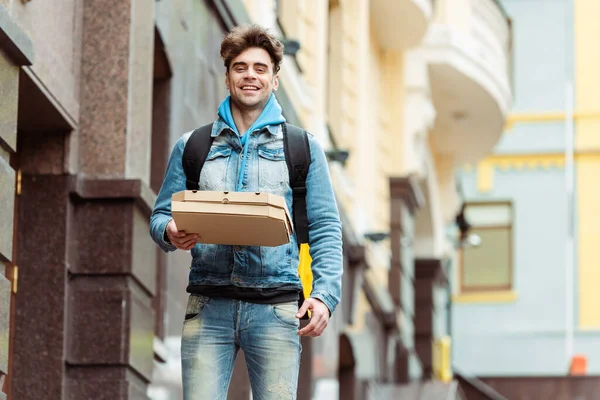 Courrier positif avec sac à dos thermique souriant à la caméra tout en tenant des boîtes à pizza dans la rue urbaine — Photo de stock