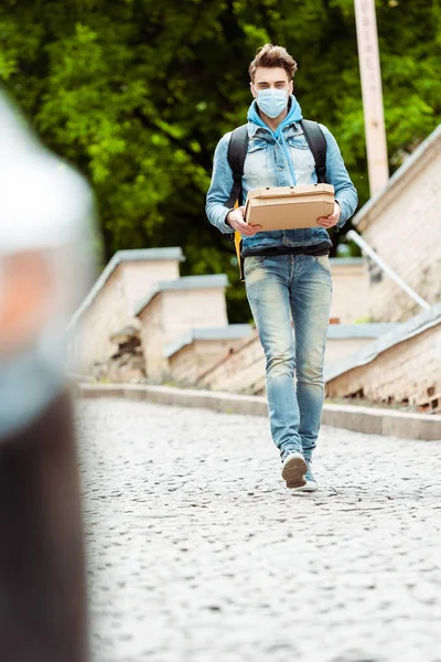 Selective focus of courier in medical mask holding pizza boxes while walking on urban street — Stock Photo