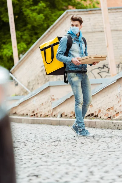 Selective focus of delivery man in medical mask with thermo backpack holding pizza boxes on urban street — Stock Photo