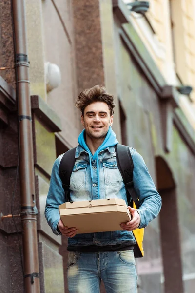 Mensajero guapo sonriendo a la cámara mientras sostiene cajas de pizza en la calle urbana - foto de stock