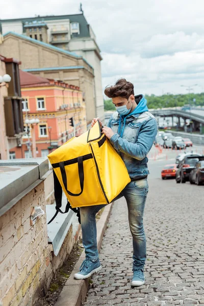 Mensajero en máscara médica sujeción bolsa de termo en la calle urbana con edificios y carretera en el fondo - foto de stock