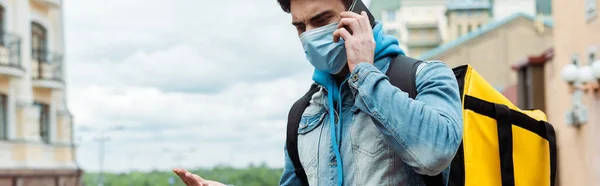 Panoramic orientation of courier in medical mask with thermo backpack pointing with hand and talking on smartphone on urban street — Stock Photo