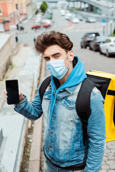 Courier in medical mask with thermo backpack showing smartphone with blank screen on urban street — Stock Photo