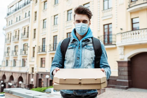 Corriere in maschera medica contenente scatole di pizza e guardando la fotocamera sulla strada della città — Foto stock