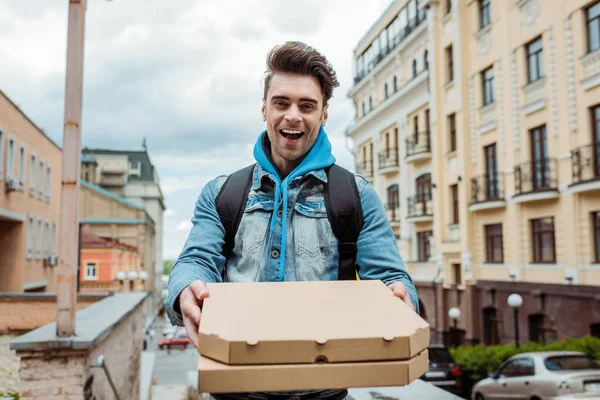 Focus selettivo del corriere allegro che tiene scatole di pizza con strada urbana sullo sfondo — Foto stock
