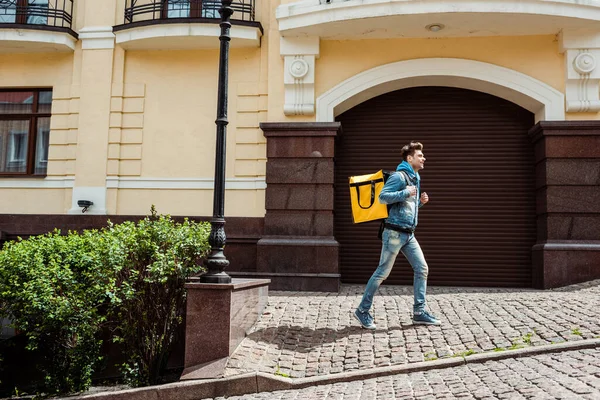 Side view of smiling courier with thermo backpack walking on paving stone walkway near buildings — Stock Photo