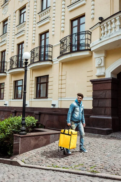 Livreur souriant tenant un sac thermique tout en marchant dans la rue urbaine — Photo de stock