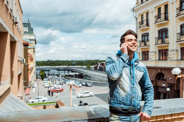 Красивый мужчина улыбается во время разговора по смартфону с облачным небом и городской улицей на заднем плане — стоковое фото