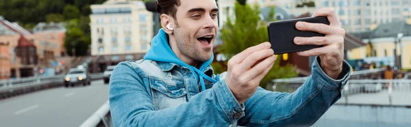 Panoramic shot of excited man in earphones taking photo with smartphone on urban street — Stock Photo
