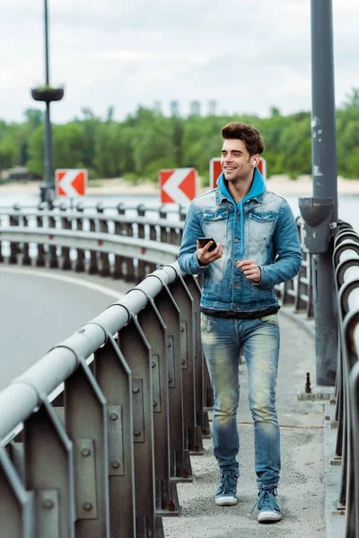 Selective focus of positive man holding cellphone while walking near railing on bridge — Stock Photo