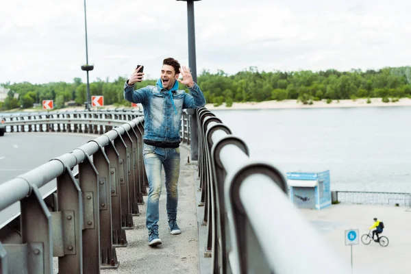 Focus selettivo dell'uomo allegro che fa videochiamate sullo smartphone mentre cammina sul ponte — Foto stock