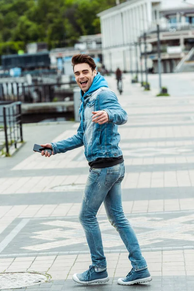 Side view of cheerful man in earphones holding smartphone on urban street — Stock Photo
