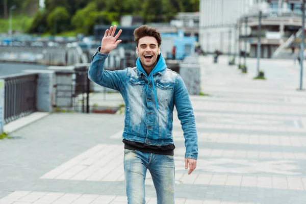 Positive man in earphones waving hand at camera on urban street — Stock Photo