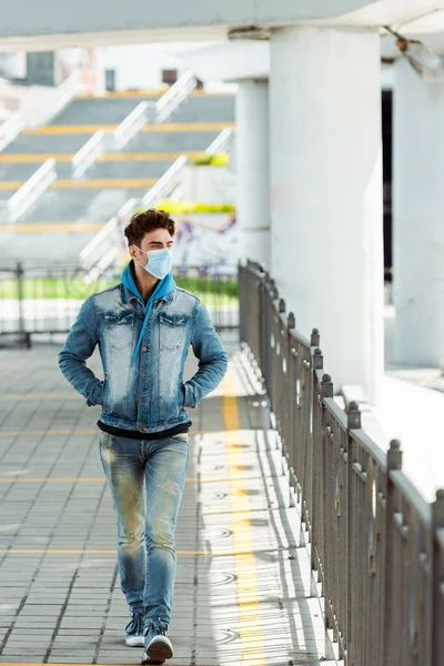 Hombre joven en máscara médica con las manos en bolsillos de chaqueta caminando por la calle urbana - foto de stock