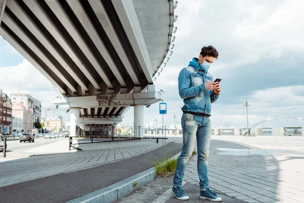 Uomo in maschera medica e giacca di denim utilizzando smartphone sulla strada urbana vicino al ponte — Foto stock