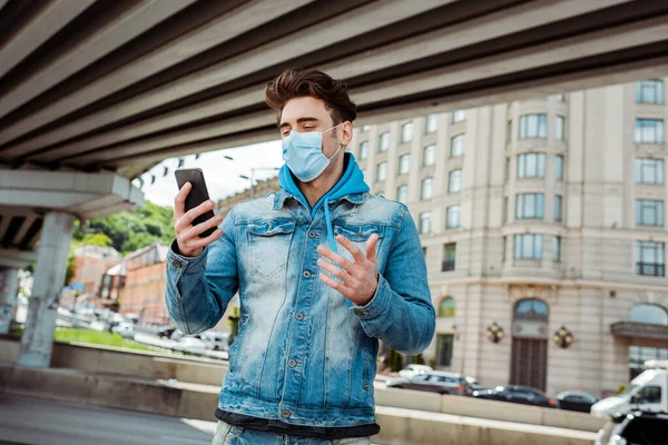 Hombre con máscara médica sosteniendo teléfono celular en la calle de la ciudad - foto de stock