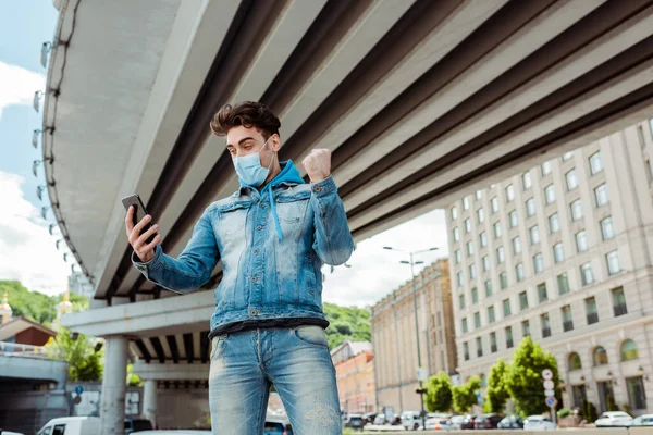 Low angle view of man in medical mask showing yeah gesture and using smartphone on urban street — Stock Photo