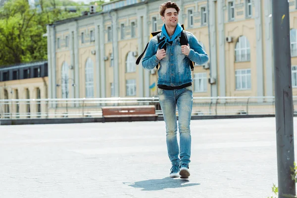 Selective focus of smiling delivery man with thermal backpack walking on urban street — Stock Photo