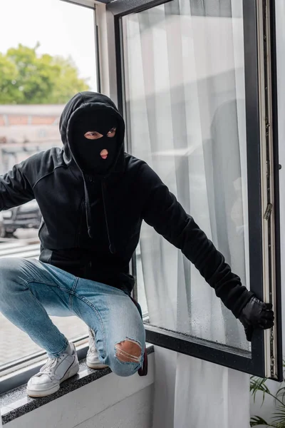 Robber in balaclava standing on open window during stealing — Stock Photo