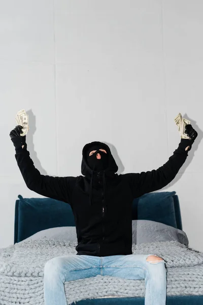 Robber in balaclava and leather gloves holding dollars on bed — Stock Photo