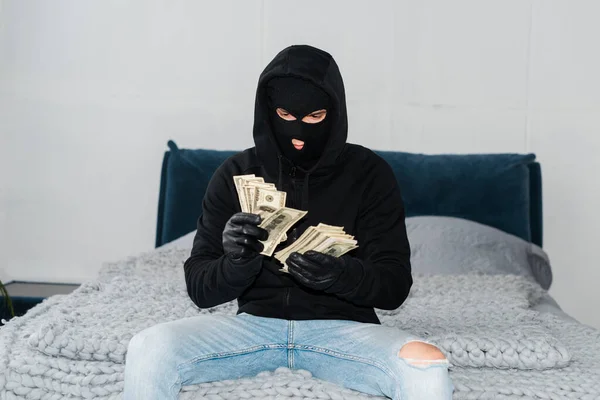 Thief in mask and leather gloves counting dollars on bed — Stock Photo