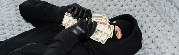 Panoramic shot of thief in balaclava holding money near face on bed — Stock Photo