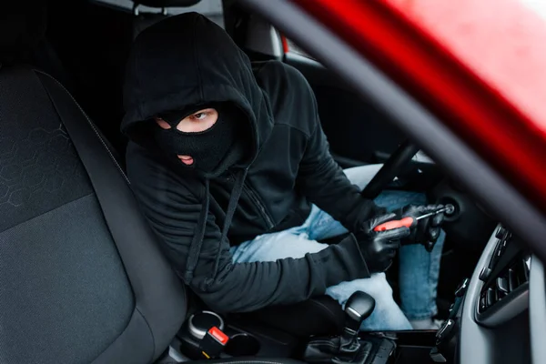 Enfoque selectivo del ladrón en pasamontañas mirando hacia otro lado mientras usa el destornillador para la cerradura de encendido en el coche - foto de stock