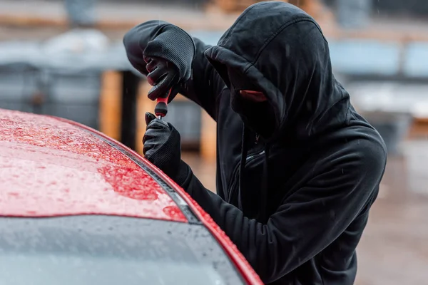 Enfoque selectivo del ladrón en máscara y guantes de cuero abriendo la puerta de auto con destornillador al aire libre - foto de stock