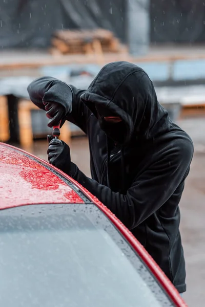 Selective focus of burglar in mask opening door of car with screwdriver during rain on urban street — Stock Photo