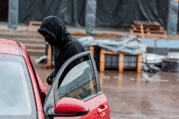 Vista laterale del ladro in passamontagna in piedi vicino all'auto con porta aperta sulla strada urbana — Foto stock