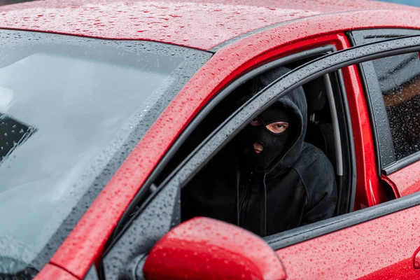 Selective focus of robber in balaclava looking at camera while sitting in car with open door — Stock Photo