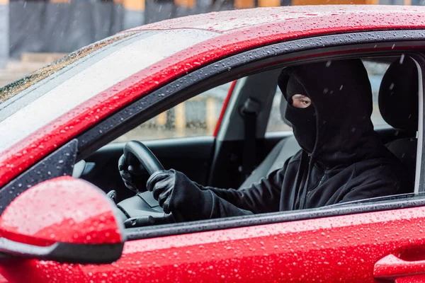 Вид сбоку грабителя в кожаных перчатках и балаклаве, сидящего в машине во время дождя — стоковое фото