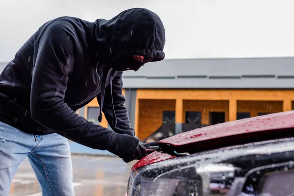 Side view of robber in balaclava holding screwdriver while opening car hood on urban street — Stock Photo